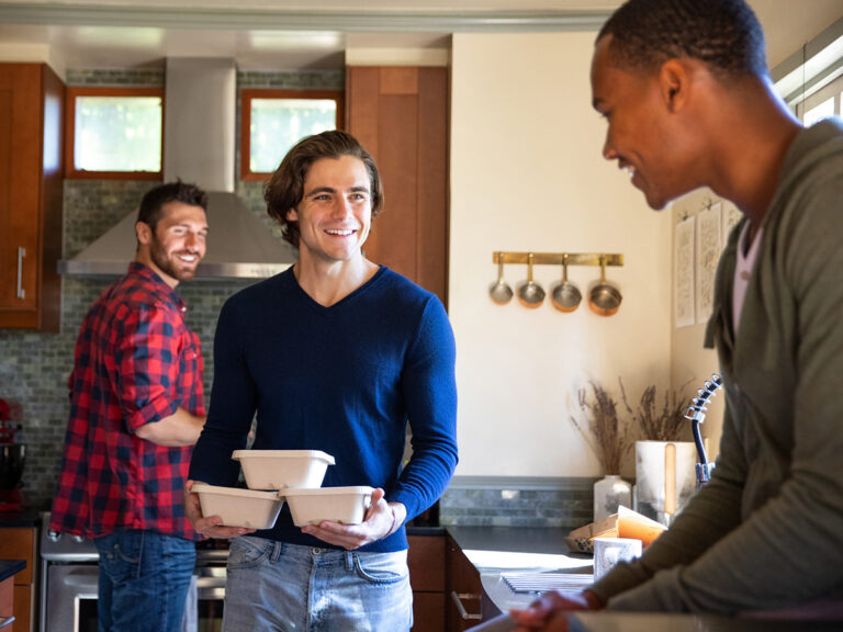 Three young men in a kitchen prepare to eat takeout food.